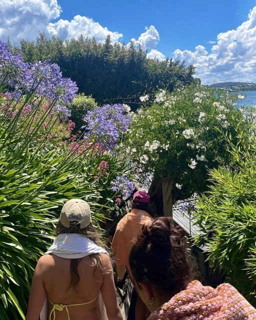 Three people walk on a path flanked by vibrant blue agapanthus flowers, green foliage, and distant blue water under a cloudy sky.