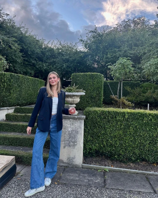 A woman in a blazer and jeans poses outside on stone steps, surrounded by green hedges and trees under a cloudy sky.