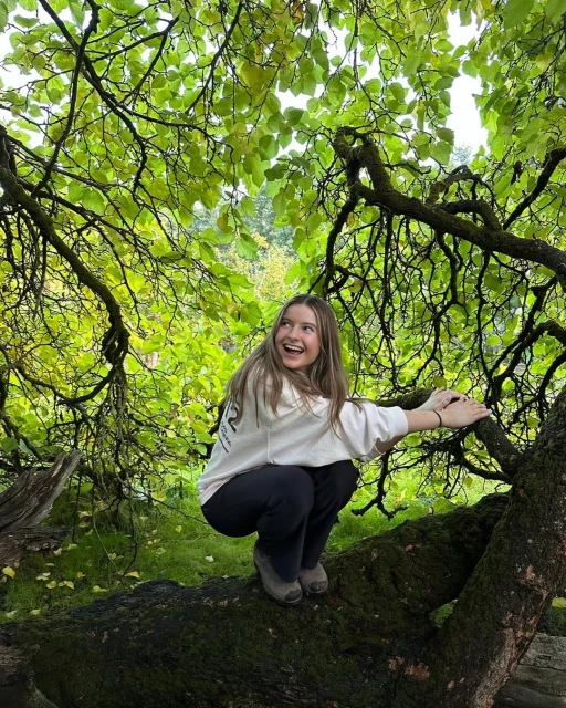Smiling young woman squats on a tree branch under a canopy of green leaves outdoors. She wears a casual sweatshirt and dark pants.