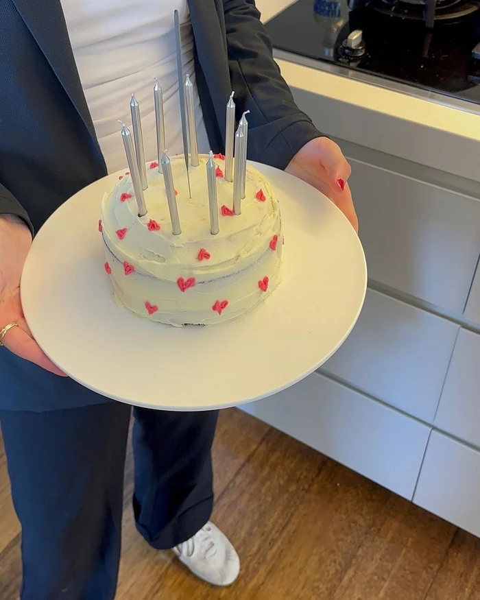 Person in a dark suit holding a white plate with a frosted cake decorated with small red hearts and tall candles.