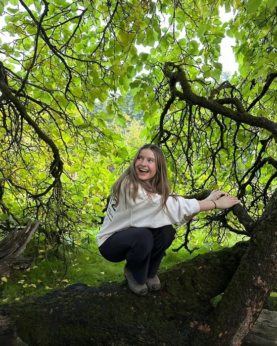 A young woman squatting on a large mossy tree branch underneath a dense canopy of vibrant green leaves, smiling.
