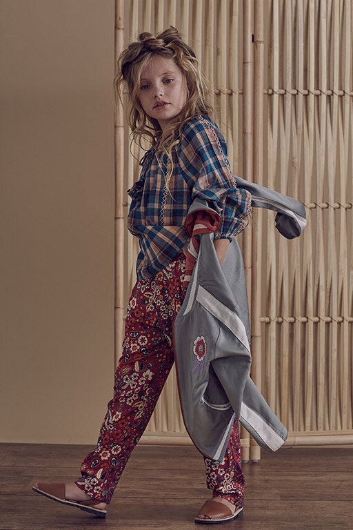 A young girl models a plaid top and floral pants, indoors. The girl is walking to the side with a jacket over her arm.