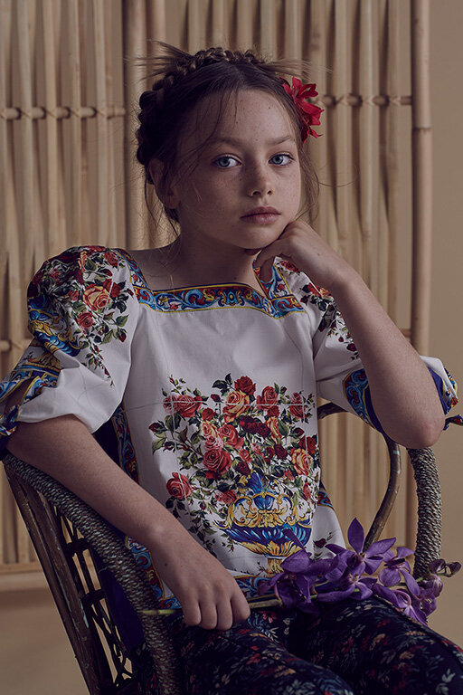 Young girl with braided hair, wearing a floral blouse, sits in a wicker chair in front of a bamboo background, holding purple flowers.