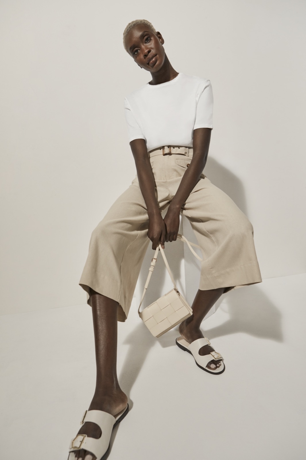 Fashion shoot of a Black woman in a white top and beige pants, sitting and holding a purse, with white heeled sandals, indoors.