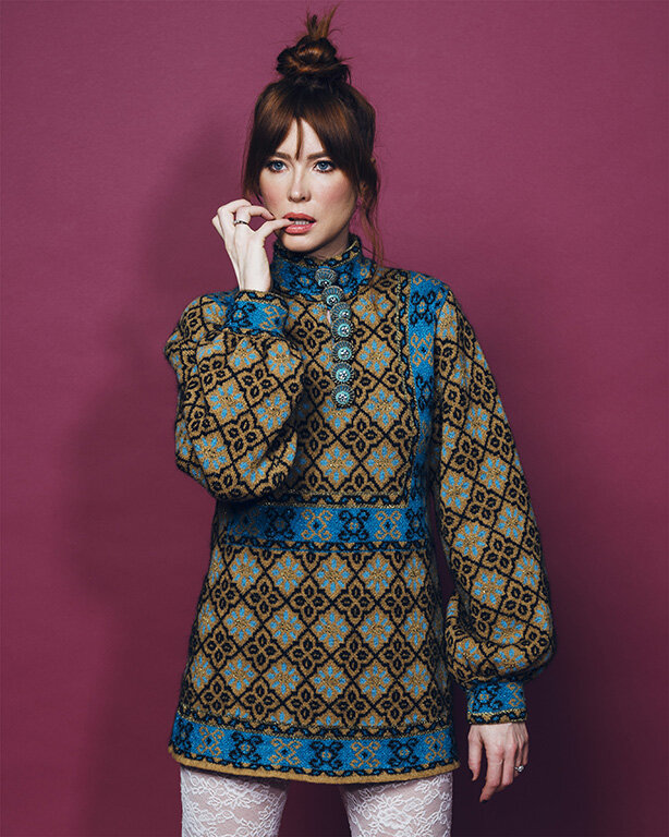 Red-haired woman in patterned tunic and white lace tights poses against a magenta background with hand near her face.
