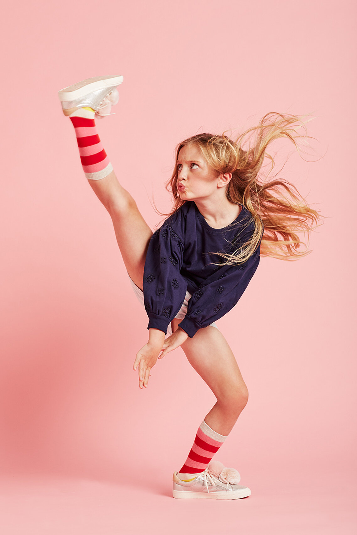 A young girl with blonde hair and wearing a dark top and pink striped socks is kicking one leg high against a pink backdrop.