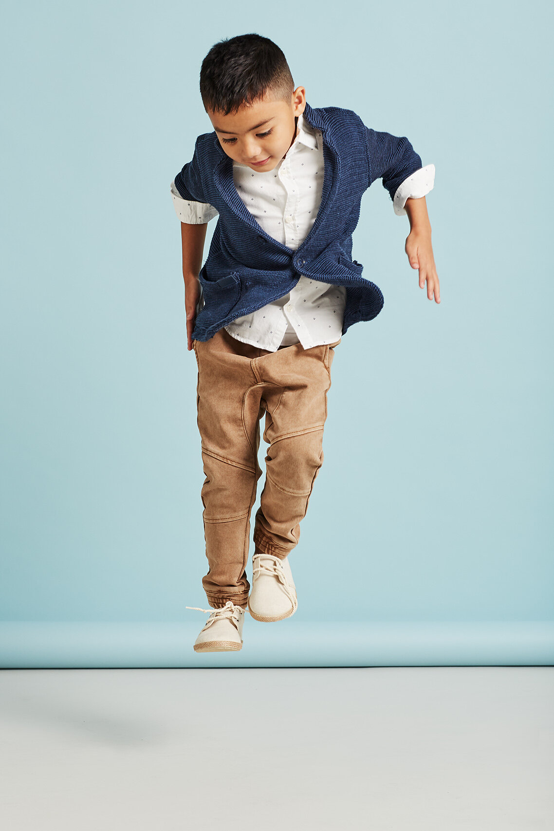 Full shot of a young boy with an open blue cardigan, white shirt, and brown pants mid-air against a light blue background in a studio.