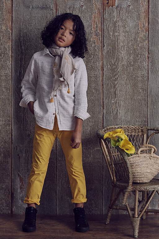 Full shot of a curly-haired child in a button-down shirt and yellow pants standing in front of a rustic wooden wall, fashion portrait.