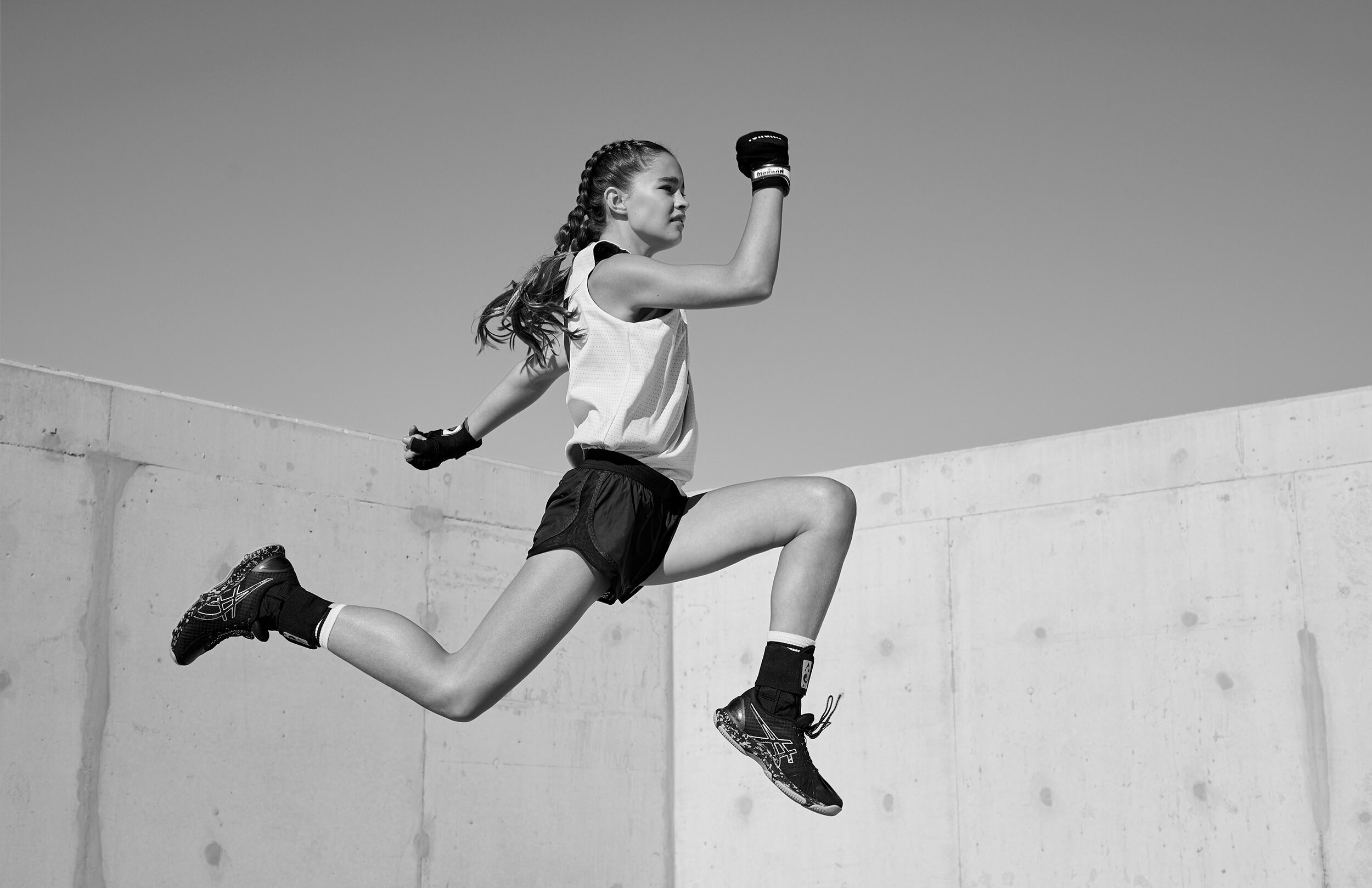 Athlete in mid-air leap, showcasing athletic apparel; black and white photo with a bright, cloudless sky and concrete wall backdrop.
