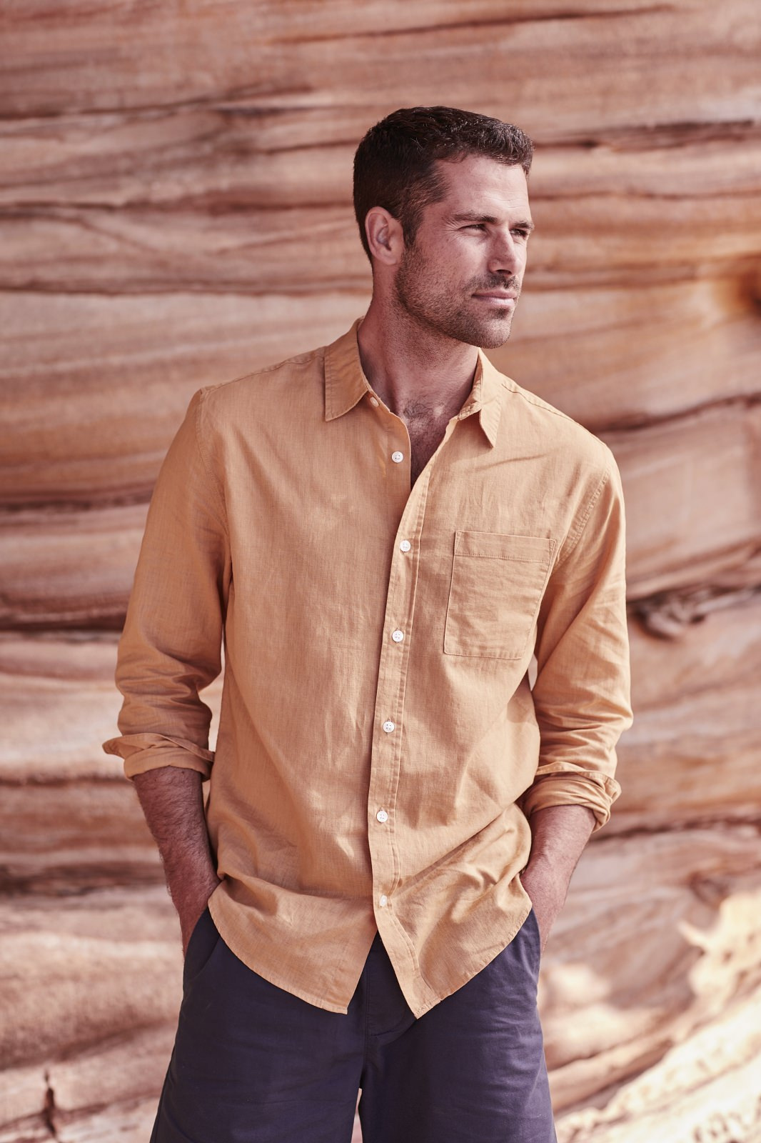 Adult male poses in front of a textured beige stone wall. He wears a golden-brown, long-sleeved button-down shirt and dark shorts.