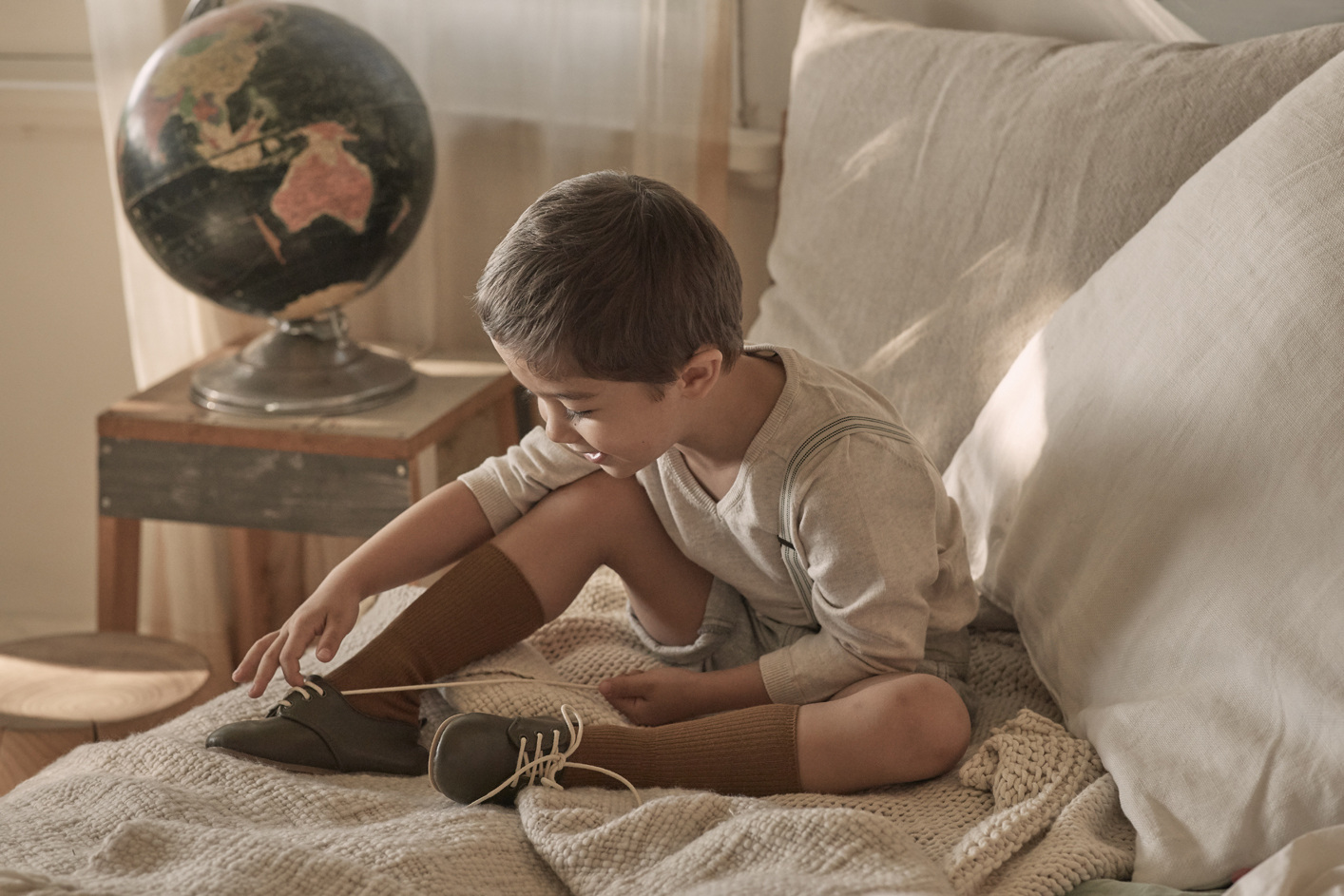 A young boy sits on a bed with a globe in the background, lacing up green shoes with brown knee-high socks in a warm, indoor setting.
