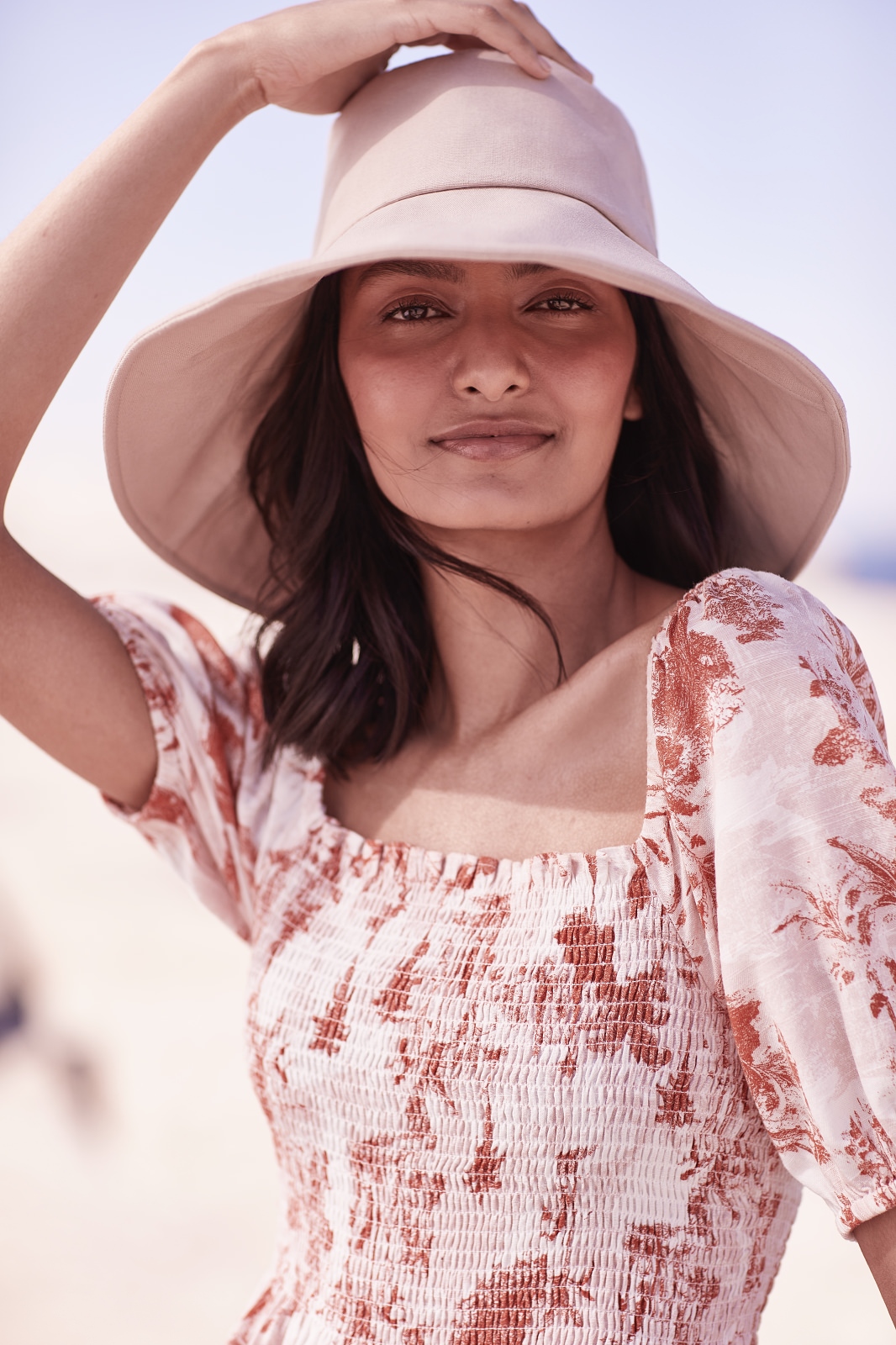 Brunette woman wearing a beige sun hat and a floral print dress poses on a beach with white sand and blue sky in soft, natural light.