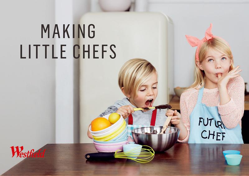 Two fair-skinned children, a boy and a girl, baking together in an indoor kitchen with kitchenware and food on a brown wooden table.