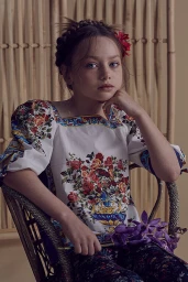 Young girl with braided hair and red floral hairpin seated in a rattan chair wearing a floral blouse by stylist Emma Wood.
