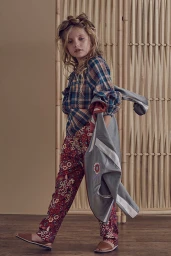 Young girl modeling a plaid blouse and patterned trousers while holding a grey jacket against a bamboo screen.