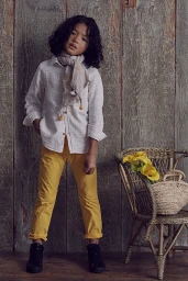 Young child with curly dark hair wearing a white button-down shirt, yellow trousers, and a patterned scarf, standing indoors.