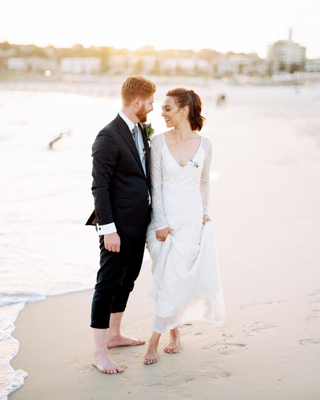 A happy barefoot couple dressed in wedding attire gazes at each other on a sandy beach with buildings visible in the distance during daytime.