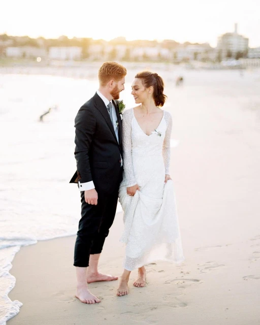 A happy barefoot couple dressed in wedding attire gazes at each other on a sandy beach with buildings visible in the distance during daytime.