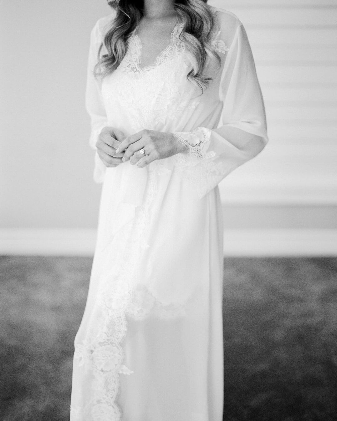 Woman wearing a white lace-trimmed robe stands indoors. Bridal fashion and delicate textile details are highlighted in a soft, neutral palette.