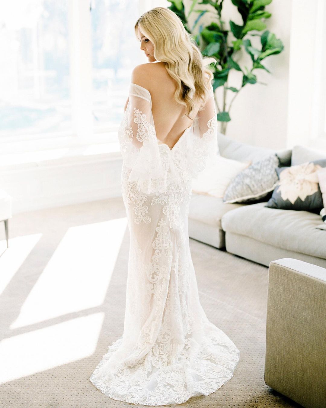 Elegant back view of a woman with blonde hair in a sheer white, lace wedding dress, posing in an airy, light-filled home interior.