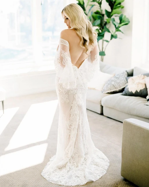 Elegant back view of a woman with blonde hair in a sheer white, lace wedding dress, posing in an airy, light-filled home interior.