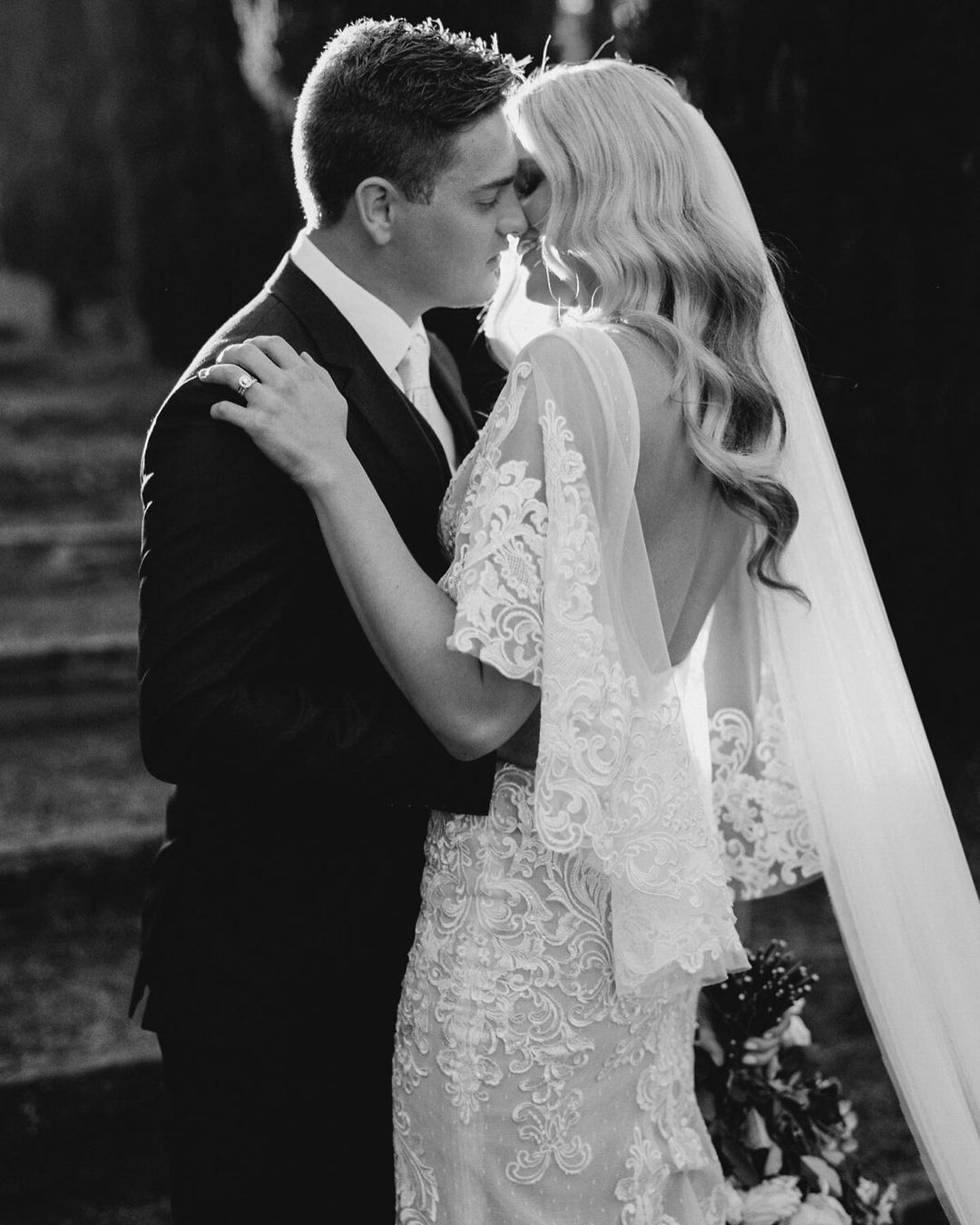 Monochrome portrait of a bride and groom embracing. The bride is in an intricate lace gown with a long veil, backlit for dramatic effect.