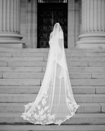 Monochromatic image captures a bride from behind, ascending stone steps towards an ornate door, her long, lace-edged veil cascading down.