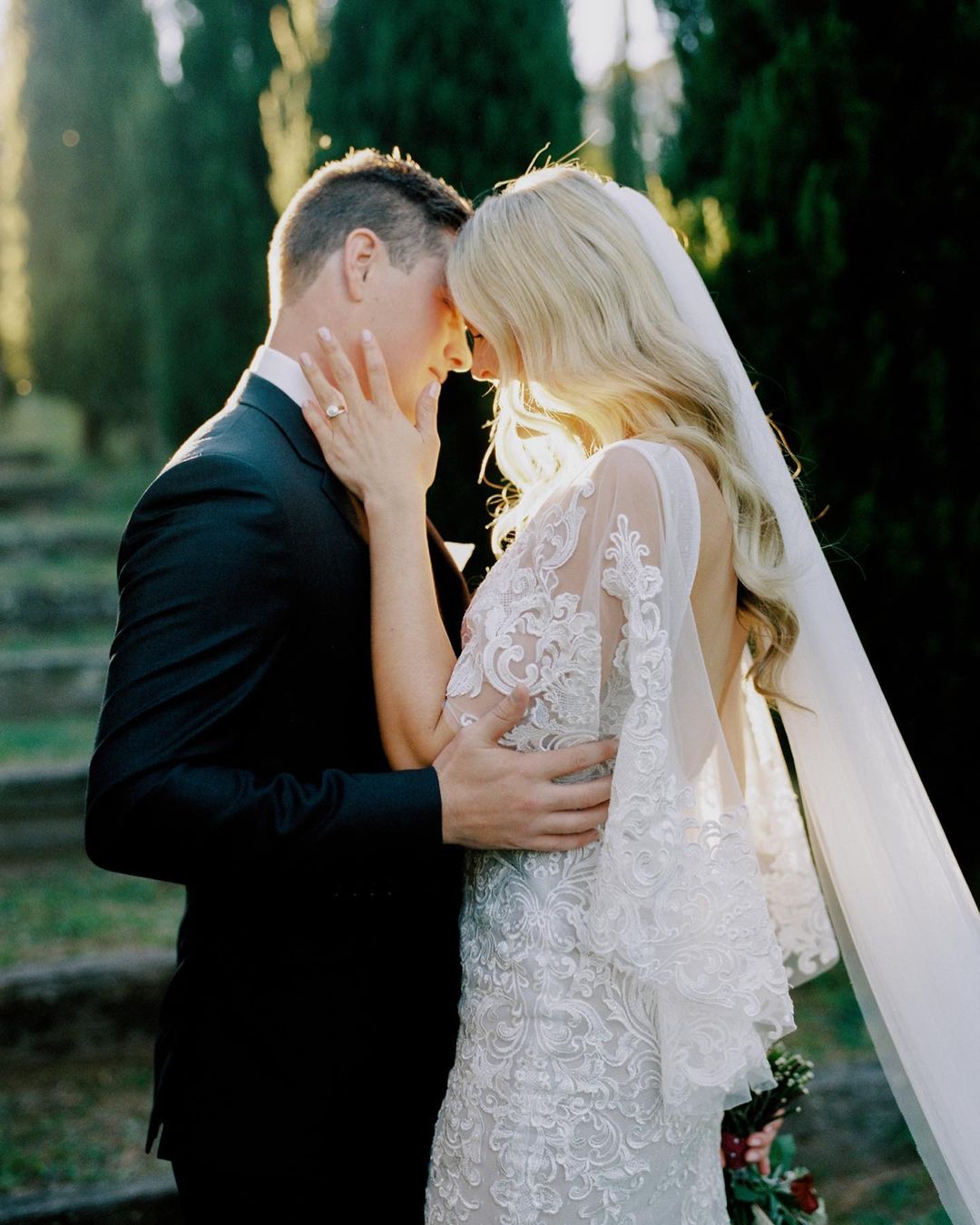 An embracing couple on their wedding day, the woman in a white, floral lace gown and veil, the man in a classic black suit. Outdoor setting.