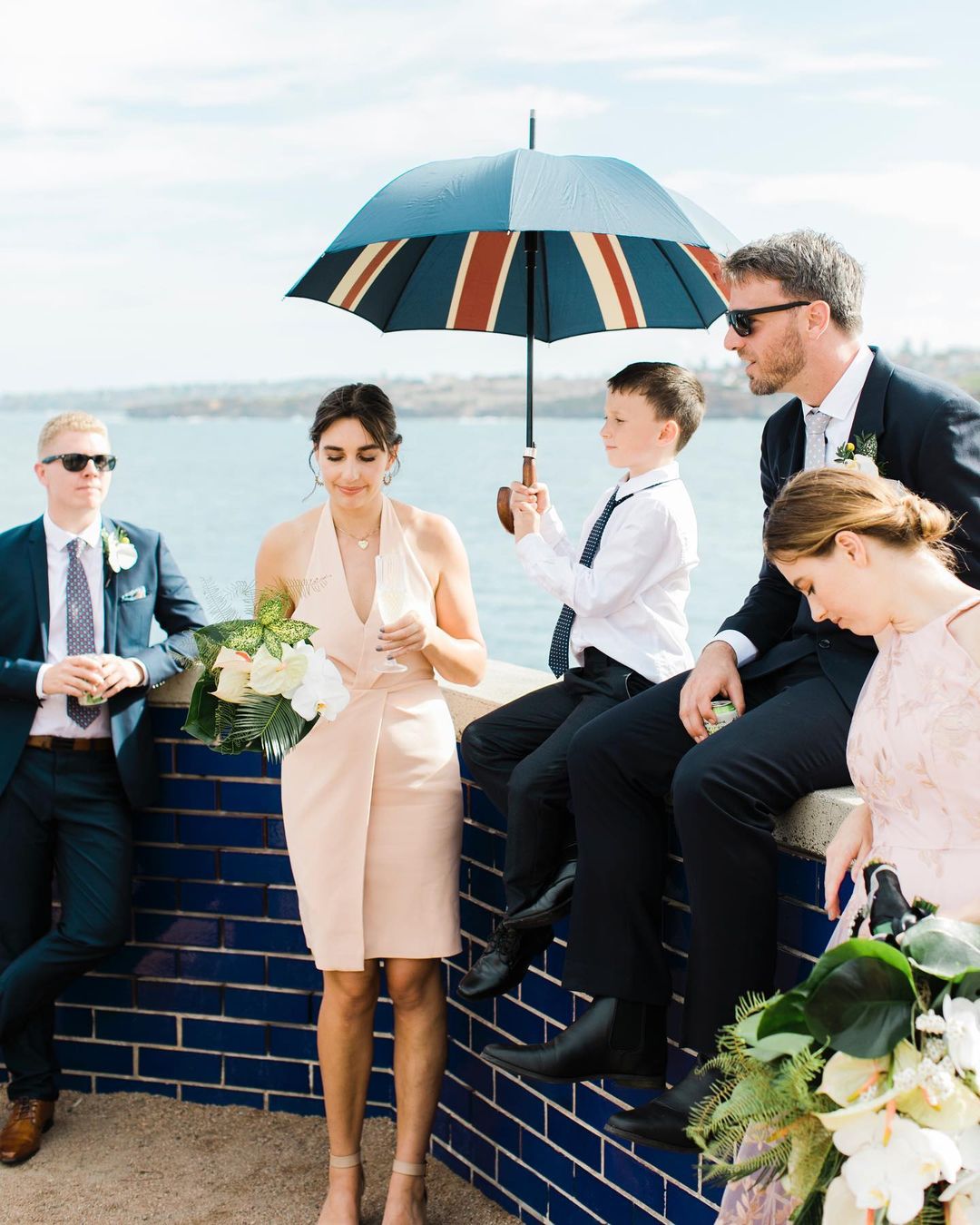 Outdoor gathering with a group of people, including men, women, and a child, on a sunny day near the ocean with an umbrella and bouquets.