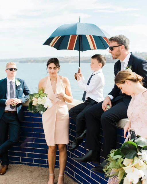 Outdoor gathering with a group of people, including men, women, and a child, on a sunny day near the ocean with an umbrella and bouquets.
