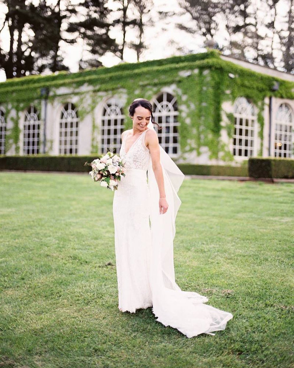 A woman in a white wedding dress smiles and poses outdoors with a flower bouquet, with a vine-covered building in the background.