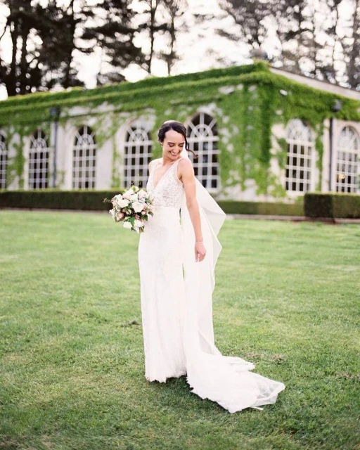 A woman in a white wedding dress smiles and poses outdoors with a flower bouquet, with a vine-covered building in the background.
