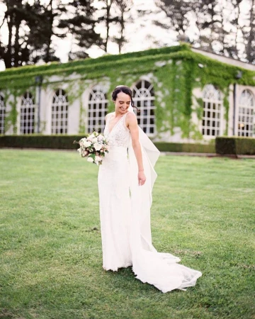 A woman in a white wedding dress smiles and poses outdoors with a flower bouquet, with a vine-covered building in the background.