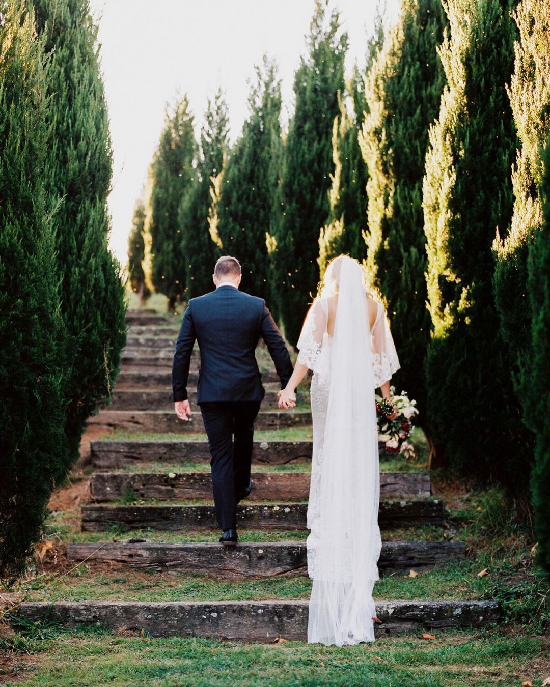 Bride and groom in wedding attire walk hand-in-hand up mossy stone steps flanked by rows of tall evergreen trees in golden light.
