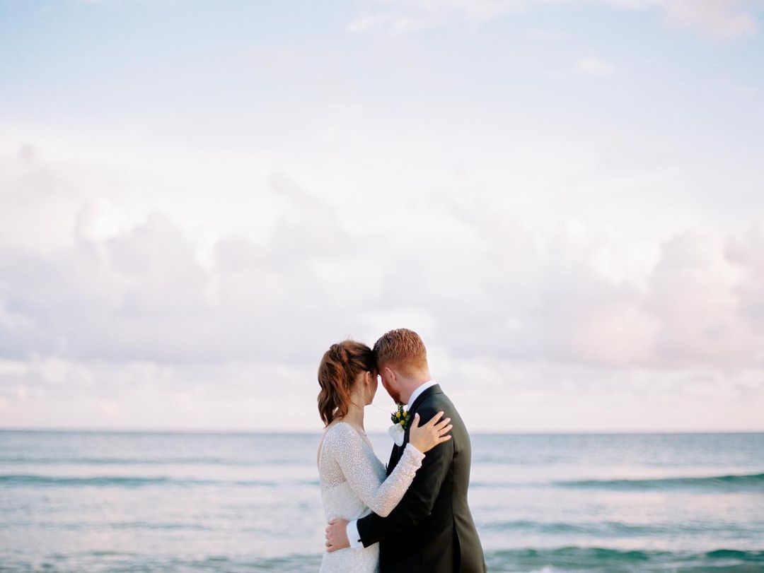 Wedding couple embraces on a beach with soft, romantic lighting. The focus is on their connection against the serene coastal backdrop.