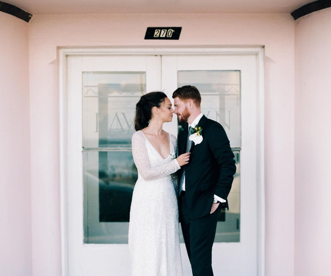 Bride and groom in wedding attire embrace in front of doors with address '270', a light-pink building, bright natural lighting, and formal wear.