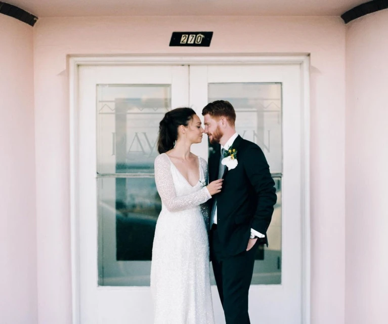 Bride and groom in wedding attire embrace in front of doors with address '270', a light-pink building, bright natural lighting, and formal wear.