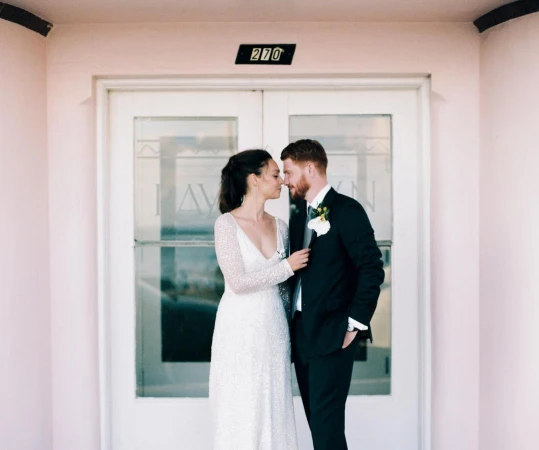 Bride and groom in wedding attire embrace in front of doors with address '270', a light-pink building, bright natural lighting, and formal wear.