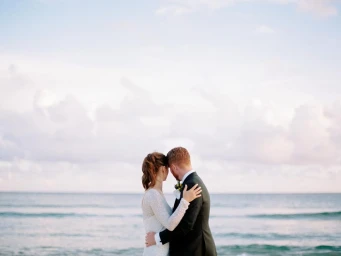 Bride in a white gown and groom in a dark suit embracing on a sandy beach by the ocean, captured from behind.