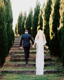 Bride and groom holding hands walking up stone garden steps lined with tall cypress trees, photographed by Glen Edwards.