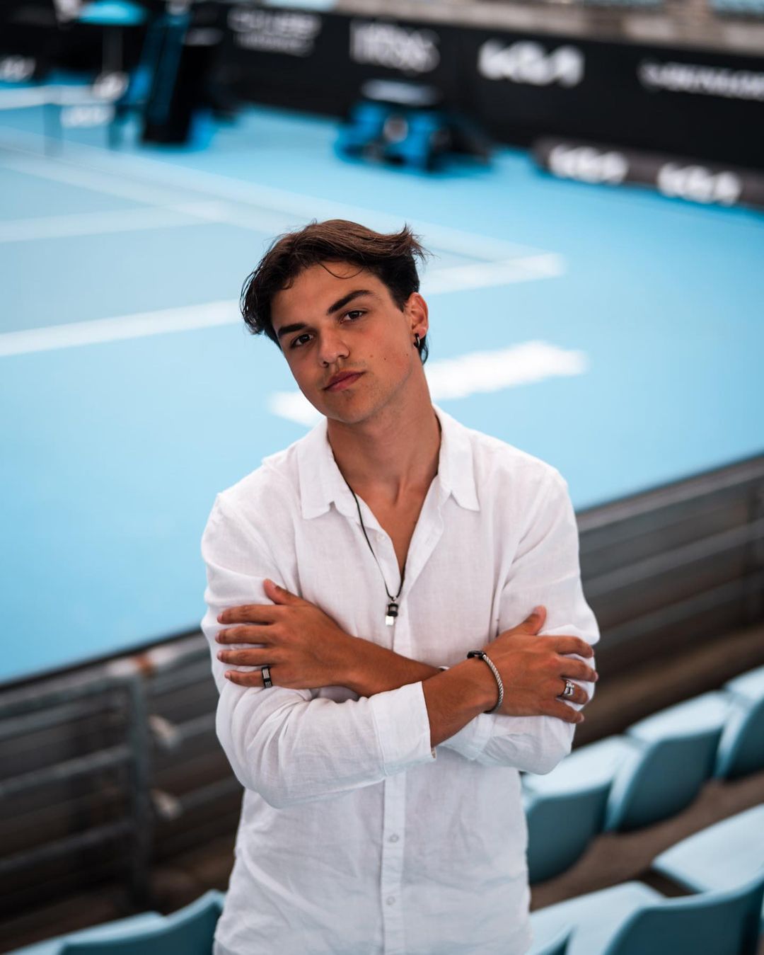 Portrait of a young adult male with arms crossed in front of a tennis court, wearing a white button-down shirt and silver jewelry.