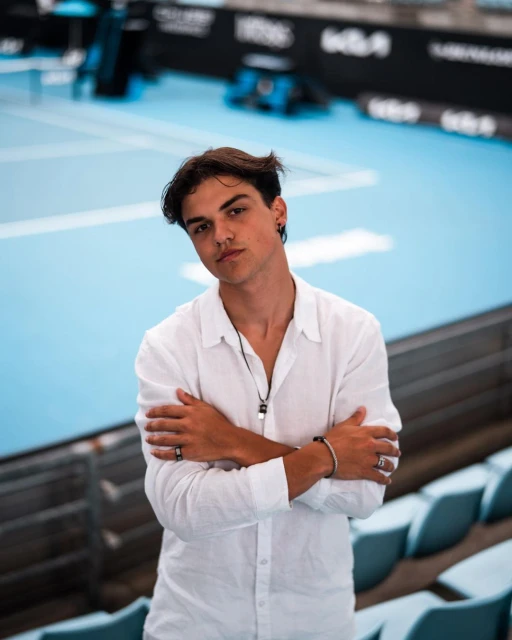 Portrait of a young adult male with arms crossed in front of a tennis court, wearing a white button-down shirt and silver jewelry.