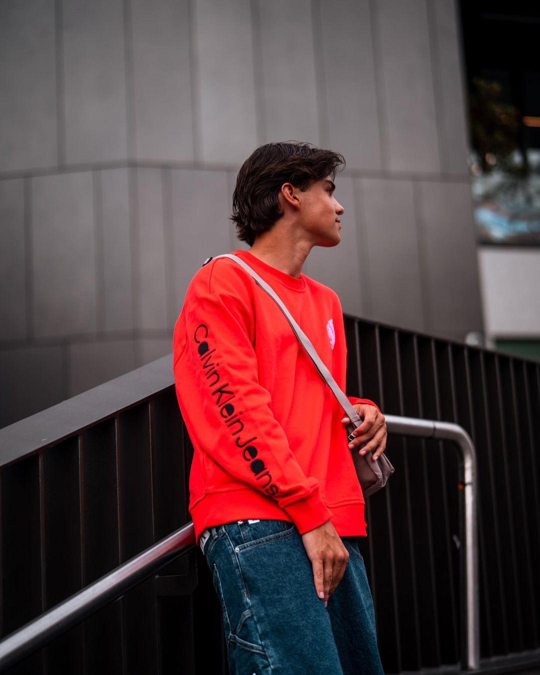 Young man in a red sweatshirt and blue jeans stands outdoors against a grey wall, looking off to the side.