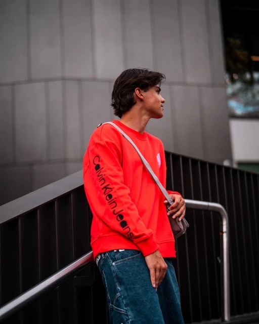Young man in a red sweatshirt and blue jeans stands outdoors against a grey wall, looking off to the side.