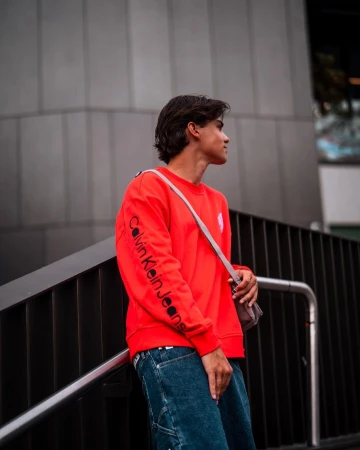 Young man in a red sweatshirt and blue jeans stands outdoors against a grey wall, looking off to the side.