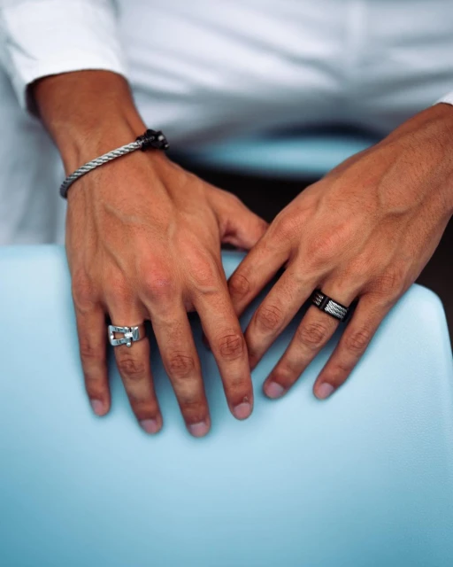 Close-up of male hands with rings and a bracelet, resting on a blue surface, showcasing jewelry details and masculine fashion.