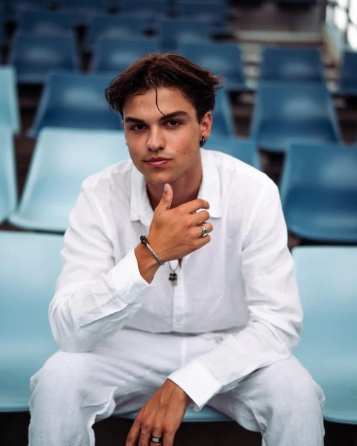 Young man in a white linen outfit poses in a stadium, seated against a backdrop of blue stadium seats, creating a casual portrait.