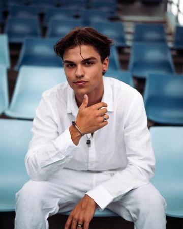 Young man in a white linen outfit poses in a stadium, seated against a backdrop of blue stadium seats, creating a casual portrait.