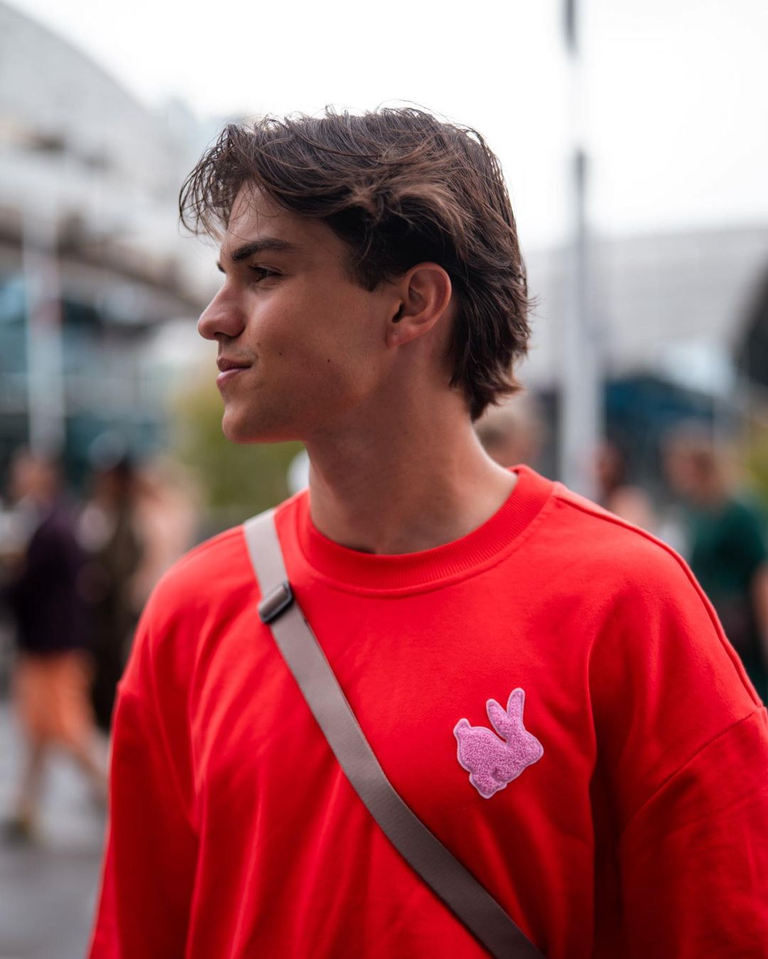 A young man in a red sweatshirt stands outdoors, looking off to the side with a gray messenger bag strap across his chest.