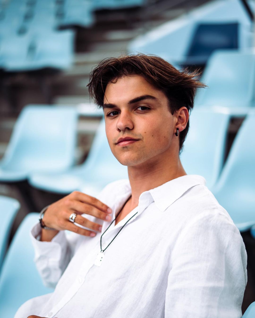 Portrait of a young man with brown hair in a white shirt seated on blue stadium seats, gazing directly at the camera with a neutral expression.